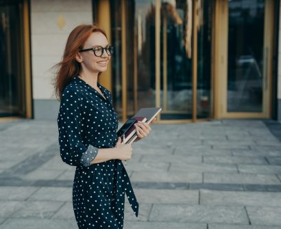 Skilled redhead businesswoman stands sideways has cheerful expression carries modern gadgets